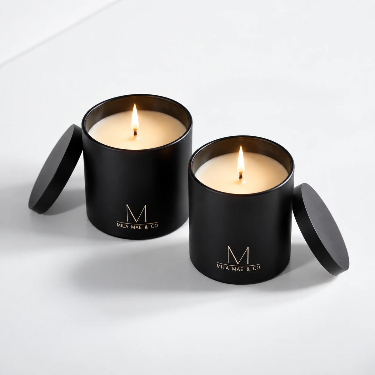 Two Mila Mae &amp; Co candles in black vessels, lit, with their lids beside them